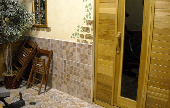 Exterior view of a sauna room with wooden panels on the walls and door. The area outside the sauna has beige tile flooring and brown square wall tiles.