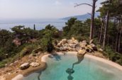 Pool with natural stone accents, offering a stunning view of the coastline and distant mountains, surrounded by trees and rocky terrain.