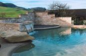 Elegant pool with a stone waterfall feature, surrounded by natural stone walls and set in a serene countryside landscape with rolling hills in the background.