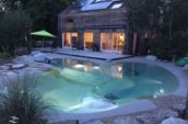 Evening view of a modern house with large glass doors opening onto a patio and a beautifully lit, organic-shaped pool surrounded by rocks and greenery.