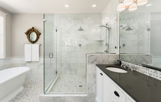 Elegant bathroom featuring a frameless glass shower enclosure with white marble walls and mosaic tile flooring. A freestanding bathtub is located near the shower, and a double sink vanity with dark countertops and a large mirror completes the space.