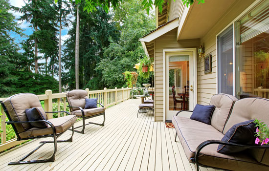 Outdoor wooden deck attached to a beige house, surrounded by lush greenery and tall trees.