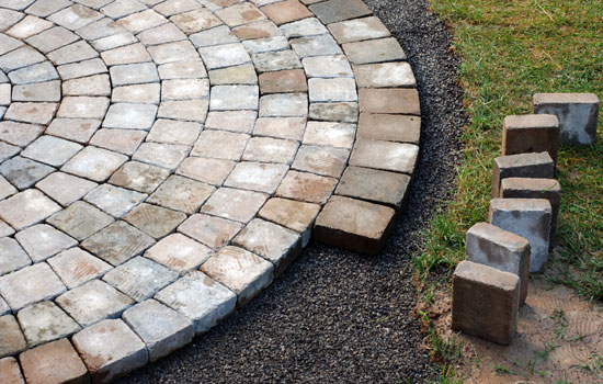 Close-up of a patio installation in progress with interlocking stone pavers being laid, bordered by standing pavers and gravel, with a grassy edge.