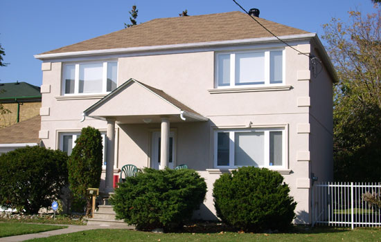 Two-story beige house with a small front porch supported by columns, surrounded by neatly trimmed bushes and a grassy lawn, photographed on a sunny day.