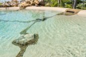Crystal-clear pool water with pebble and stone details, highlighting a rock embedded in the pool floor, surrounded by natural stone accents and greenery.
