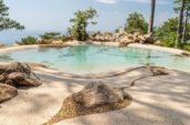 Close-up of a pool with natural stone and pebble accents, surrounded by rocks and greenery, offering a peaceful view of a body of water in the background.