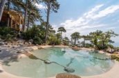 Tranquil outdoor pool with a natural stone design, surrounded by pine trees and rocky landscape, offering scenic views of the coastline and sky.