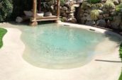 Tropical-style pool with a thatched-roof cabana and natural rock wall backdrop, surrounded by lush greenery and featuring a shallow, beach-like entry.