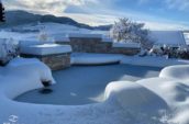 Snow-covered outdoor pool and surrounding landscape, with a stone waterfall feature, set against a backdrop of rolling hills and a bright, wintery sky.