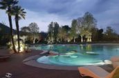 Beautifully lit outdoor pool at dusk, surrounded by palm trees and lush greenery, with soft lighting highlighting the water and landscape features.
